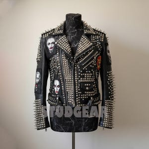 May include: Black leather jacket heavily embellished with silver studs and spikes. The jacket features multiple patches with graphic designs and a zipper closure. The word "STUDGEAR" is visible on the bottom of the jacket.