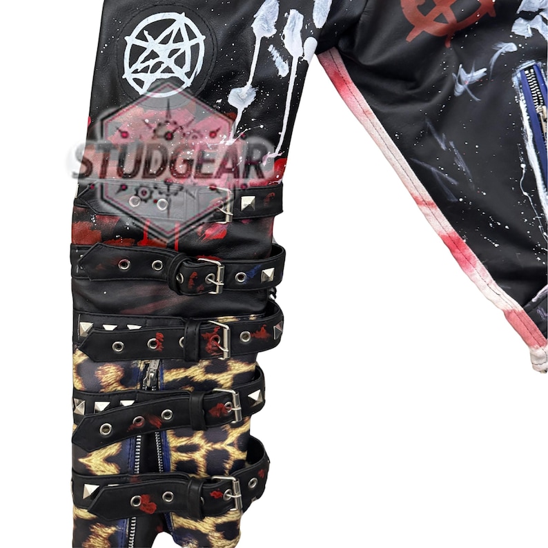 May include: A black jacket sleeve featuring white paint splatters, a pentagram symbol, and multiple black buckled straps with silver-tone studs. The lower portion displays a leopard print pattern and a vertical zip. The text STUDGEAR is visible.