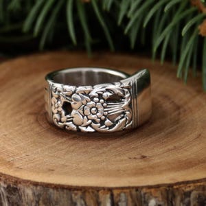 Puede incluir: Un anillo de plata hecho de una cuchara, con intrincados diseños florales y de hojas. El diseño del anillo incorpora el mango y el cuenco de la cuchara, creando una pieza de joyería única y reciclada. El anillo se muestra sobre una superficie de madera.