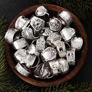 May include: A collection of silver rings, each crafted from repurposed silverware, displayed in a wooden bowl. The rings feature various floral and ornate designs, showcasing a vintage aesthetic. The arrangement is set against a dark background with evergreen sprigs.