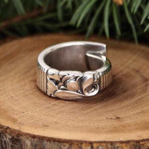 May include: A silver ring with a floral design. The ring has a wide band with vertical lines on the sides and a detailed floral pattern in the center. The ring is set on a wooden surface.