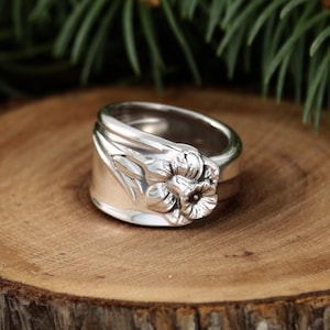 May include: A silver ring with a floral design. The ring features a detailed flower with petals and a central stamen. The ring is set on a wooden surface, with green foliage in the background.