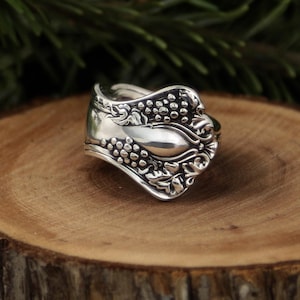 May include: A silver spoon ring with a detailed floral and grape design. The ring is set on a wooden surface, showcasing the intricate craftsmanship and vintage aesthetic. The ring's design includes a spoon bowl and a decorative handle.