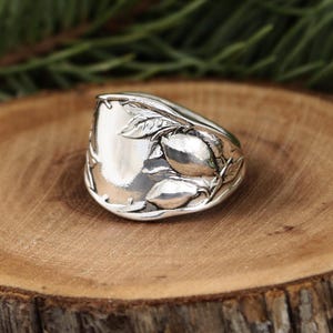 May include: A silver spoon ring with a floral design. The ring features a polished, rectangular face with detailed leaves and fruit on the sides. The ring is set on a wooden surface.