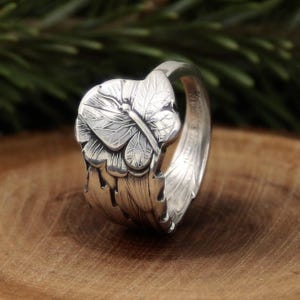 May include: A silver ring featuring a detailed butterfly design. The ring's band has a textured, leaf-like pattern, and the butterfly is centered on a floral element. The ring is displayed on a wooden surface.