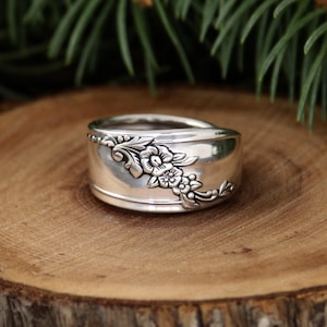 Puede incluir: Un anillo ancho de plata con un diseño floral. El anillo presenta una banda con un patrón floral detallado en la parte delantera. El anillo está sobre una superficie de madera, con agujas de pino verdes en el fondo.