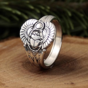 May include: A silver ring with an Egyptian scarab beetle design. The ring features a detailed beetle with outstretched wings, a sun disc, and intricate floral patterns. The ring is set against a wooden surface.