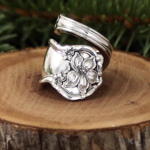 May include: A handcrafted silver spoon ring featuring a floral design. The ring is made from a repurposed spoon, with the bowl of the spoon forming the face of the ring, and the handle curving around to create the band. The ring is set on a wooden surface.