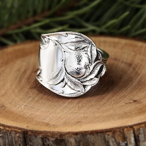 May include: A silver spoon ring with a detailed design. The ring features a central orb surrounded by leaves and floral patterns. The ring is set on a wooden surface.