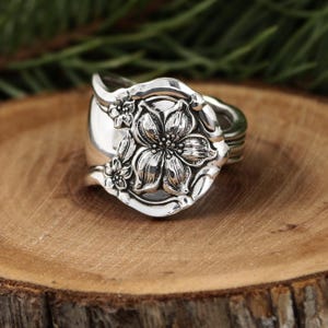 May include: A silver ring with a floral design. The ring features a large, detailed flower in the center, surrounded by smaller flowers. The ring is made from a spoon and has a vintage, handcrafted look.