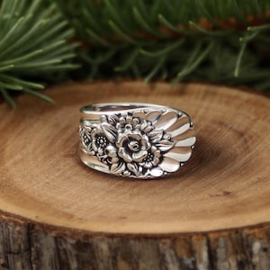 May include: A silver ring with a floral design. The ring has a detailed pattern of flowers and leaves, with a spoon-like shape. The ring is set on a wooden surface, with a blurred green background.