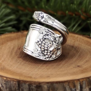 Könnte beinhalten: Ein silberner Löffelring mit einem Traubenmuster. Der Ring besteht aus einem Löffel und zeigt eine detaillierte Traube auf der Vorderseite. Das Band des Rings ist ebenfalls mit Trauben verziert.