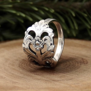 Op de afbeelding: Een zilveren ring met een sierlijk bloemmotief. De ring heeft een centrale bloem omringd door gedetailleerde bladeren en krullen. De ring ligt op een houten oppervlak, met een vage groene achtergrond.