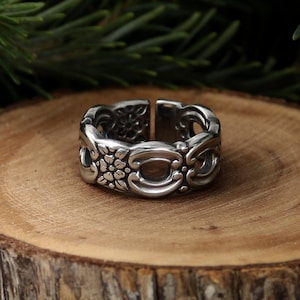 May include: A silver-colored ring with an open design. The ring features floral and scrollwork patterns. The ring is set on a wooden surface with a blurred green background.