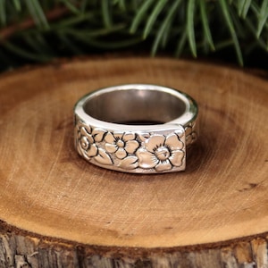 May include: A silver ring with a floral design. The ring features detailed carvings of flowers and leaves around the band. The ring is set on a wooden surface.