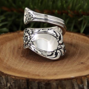 May include: A silver spoon ring with ornate floral and scroll detailing. The ring features a curved design, with the spoon's bowl forming the face of the ring. The ring is set against a wooden surface.