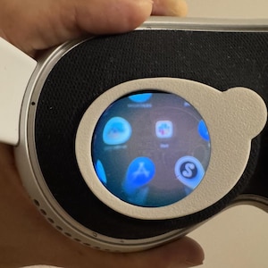 Puede incluir: Primer plano de unos auriculares de realidad virtual, con una pantalla que muestra iconos de aplicaciones. Los auriculares tienen un marco blanco y plateado con una zona acolchada negra. La pantalla está rodeada por un borde gris claro.