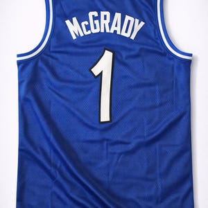 Puede incluir: Una camiseta de baloncesto azul con el nombre "McGRADY" en letras blancas en bloque sobre el número "1". El número es blanco con un contorno negro. La camiseta tiene ribetes blancos y azules alrededor del cuello y las sisas.