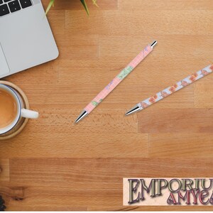 May include: Two patterned pens lie on a wooden desk, alongside a laptop and a coffee cup. One pen features a pink and green design, the other an orange and brown pattern. Both pens have silver tips. The logo "Emporium Amycat" is visible at the bottom of the image.