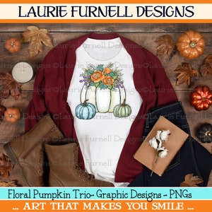 May include: A white t-shirt with a graphic design of three pumpkins with fall floral accents. The pumpkins are white, teal, and green. The text "Floral Pumpkin Trio - Graphic Designs - PNGs" is at the bottom of the image. The text "... ART THAT MAKES YOU SMILE ..." is below the first line of text.