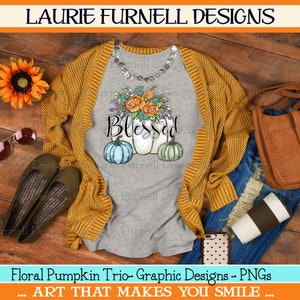 May include: A gray t-shirt with a watercolor design of three pumpkins with floral accents and the word "Blessed" in a handwritten font. The pumpkins are painted in shades of blue, green, and white. The t-shirt is displayed on a wooden surface with a brown cardigan, blue jeans, a brown leather bag, sunglasses, and a coffee cup.