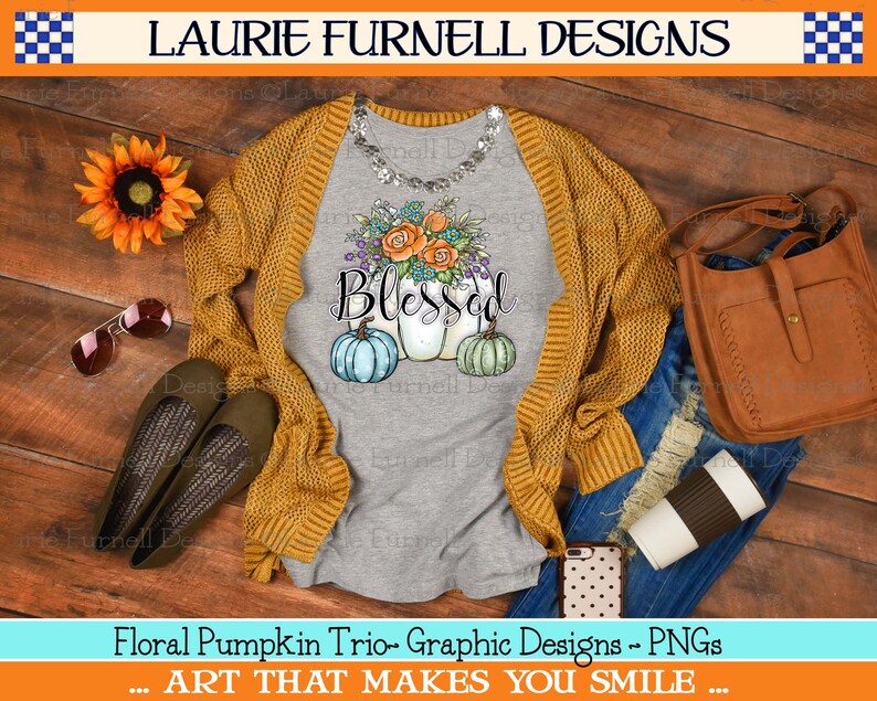 May include: A gray t-shirt with a watercolor design of three pumpkins with floral accents and the word "Blessed" in a handwritten font. The pumpkins are painted in shades of blue, green, and white. The t-shirt is displayed on a wooden surface with a brown cardigan, blue jeans, a brown leather bag, sunglasses, and a coffee cup.