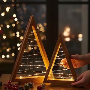 Peut inclure: Deux décorations en forme de sapin de Noël en bois avec des guirlandes lumineuses blanc chaud. Les cadres triangulaires sont en bois brun clair. De petits objets décoratifs comme des pommes de pin et des baies sont sur la table.