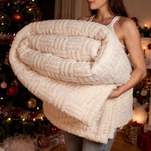 Peut inclure: Une couverture en peluche crème avec un motif texturé et côtelé. La couverture est pliée et tenue, mettant en valeur son aspect doux et invitant. L'arrière-plan comprend un sapin de Noël décoré et des décorations festives.