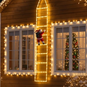 Puede incluir: Decoración navideña festiva con un Papá Noel trepando por una escalera iluminada en una casa. La casa tiene luces de cadena alrededor de las ventanas y el borde del techo, con un árbol de Navidad visible a través de una ventana.