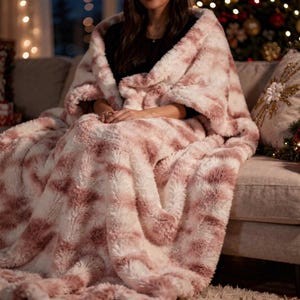 Peut inclure: Une couverture en fausse fourrure rose et blanche, drapée sur une personne assise sur un canapé. La couverture est moelleuse et crée une ambiance chaleureuse. Un sapin de Noël et un coussin décoratif sont visibles en arrière-plan.