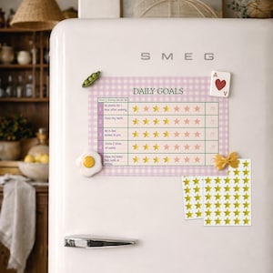 May include: A white SMEG refrigerator with a magnetic daily goals chart, star stickers, and decorative magnets. The chart has a pink gingham background and lists daily tasks with star ratings. Other magnets include a playing card and a fried egg.