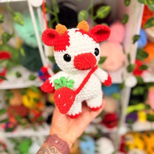 May include: A handmade crochet cow plushie with a red and white color scheme. The cow has black eyes, red ears, and a red nose. It carries a red strawberry bag. The plushie is held in a person's hand.