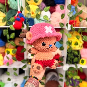 May include: A handmade crochet plush toy of a cartoon character, featuring a pink hat with a white cross, brown antlers, and red shorts. The toy is held in front of a shelf filled with colorful yarn balls and decorative greenery.