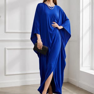 May include: A long, flowing cobalt blue dress with wide sleeves and a high side slit. The dress is paired with gold heels and a black clutch. The model is also wearing a pearl necklace and bracelet. The dress has a relaxed, elegant style.