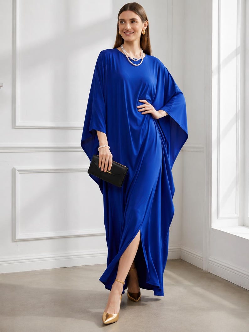 May include: A long, flowing cobalt blue dress with wide sleeves and a high side slit. The dress is paired with gold heels and a black clutch. The model is also wearing a pearl necklace and bracelet. The dress has a relaxed, elegant style.