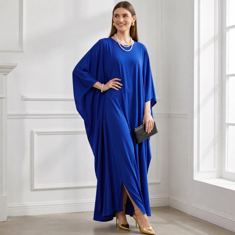 May include: A long, flowing cobalt blue dress with wide sleeves and a side slit. The dress is paired with gold heels and a small black clutch. The model is also wearing a pearl necklace and bracelet.