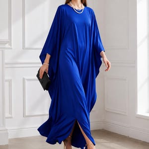 May include: A flowing, cobalt blue caftan dress with wide sleeves and a high side slit. The dress is paired with gold-colored high heels and a black clutch. The model is also wearing a pearl necklace and earrings.