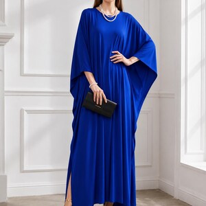 May include: A long, flowing cobalt blue dress with wide sleeves and a high side slit. The dress is accessorized with a pearl necklace, bracelet, and gold heels. A black clutch is held in the hand.