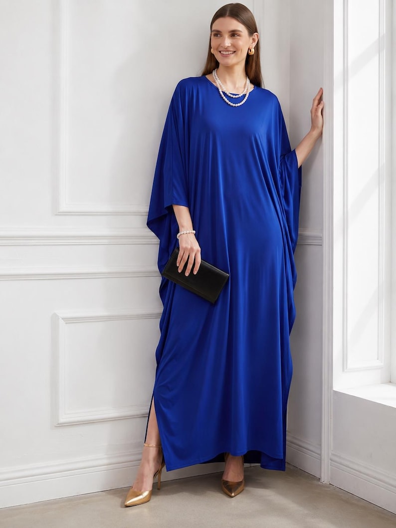 May include: A long, flowing cobalt blue dress with wide sleeves. The dress is paired with gold heels and a black clutch. The model is wearing a pearl necklace and bracelet. The dress has a slit on the side.