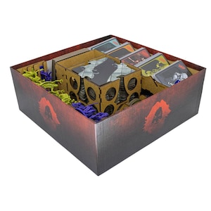 May include: A square board game box with a dark red and black gradient exterior featuring a red silhouette logo. Inside are wooden dividers, stacks of rectangular playing cards with D&D branding, and small plastic miniature figures in lime green and purple.