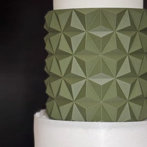 Könnte beinhalten: Mehrstöckige Torte mit geometrischem Design. Die mittlere Ebene weist ein strukturiertes, olivgrünes Muster aus sich wiederholenden Dreiecksformen auf. Die obere und untere Ebene sind glatt und weiß, was einen Kontrast erzeugt. Der Hintergrund ist schwarz.