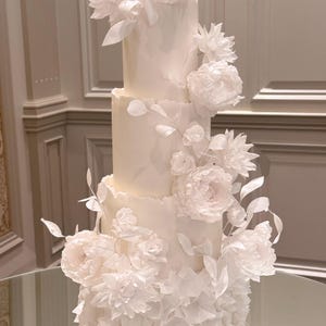 Könnte beinhalten: Eine vierstöckige weiße Hochzeitstorte, verziert mit großen, zarten weißen Zuckerblumen und Blättern. Die Torte hat einen gerüschten Boden und wird auf einer verspiegelten Oberfläche präsentiert, was eine elegante Präsentation ergibt.