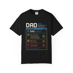 Puede incluir: Una camiseta negra de manga corta con un estampado gráfico que imita una pantalla de actualización de software. El texto dice DAD MODE INSTALLING, PATCH NOTES, DAD v3.2.7, NEW FEATURES, BUG FIXES, KNOWN ISSUES y APPLYING UPDATES con iconos de parrilla y café.