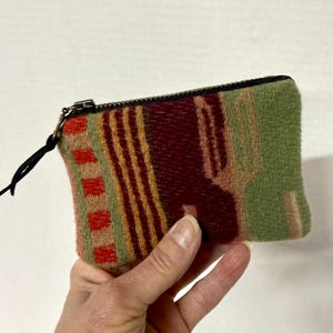 May include: A small, rectangular coin purse with a colorful woven pattern. The purse features a black zipper and a black pull tab. The pattern includes red, green, and brown vertical stripes and geometric shapes. The purse is held in a hand.