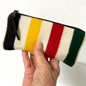 May include: A rectangular zippered pouch made of a textured off-white fabric featuring vertical stripes in black, yellow, red, and dark green. The pouch includes a black zipper closure with a black leather pull tab attached to the metal slider.