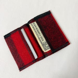 May include: Open red and black plaid wallet with a black trim. The wallet contains a one-dollar bill and a silver credit card. The wallet is made of a soft, felt-like material.