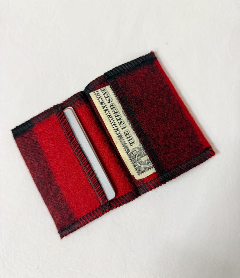 May include: Open red and black plaid wallet with a black trim. The wallet contains a one-dollar bill and a silver credit card. The wallet is made of a soft, felt-like material.