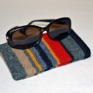 May include: Black sunglasses with a tortoiseshell pattern rest on a colorful, striped glasses case. The case features vertical stripes of red, blue, gray, tan, and yellow. The case is rectangular and made of a textured material.