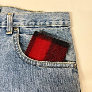 May include: A red and black plaid wallet peeks out of the front pocket of a pair of light blue denim jeans. The wallet's pattern is a classic buffalo check, and the jeans have a classic five-pocket design.