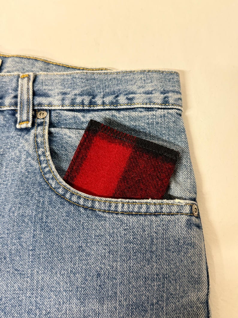 May include: A red and black plaid wallet peeks out of the front pocket of a pair of light blue denim jeans. The wallet's pattern is a classic buffalo check, and the jeans have a classic five-pocket design.